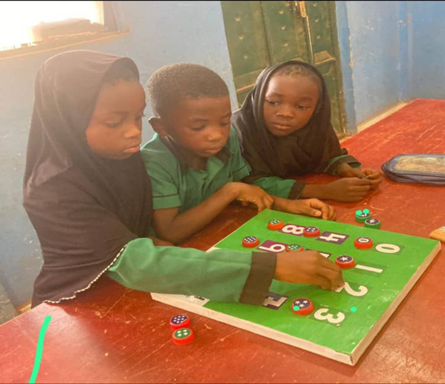 Three young students in green uniforms working together on a green learning board with numbered counting activities using colorful tokens